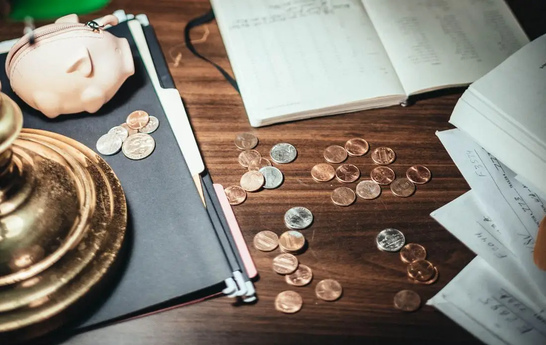 Coins and a ceramic piggy bank on a wooden desk beside an open notebook used for tracking spending