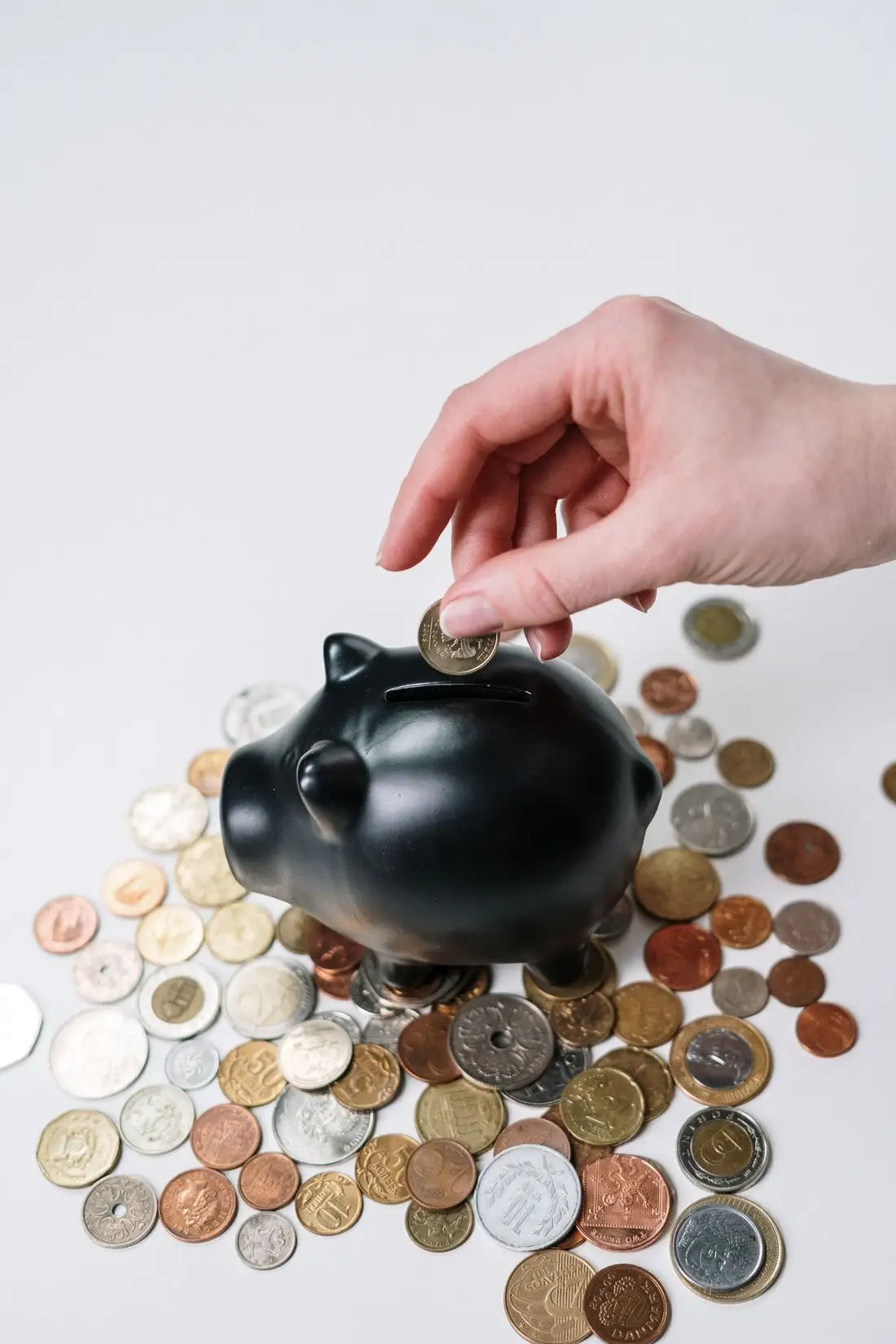 A hand dropping a coin into a black piggy bank surrounded by scattered coins