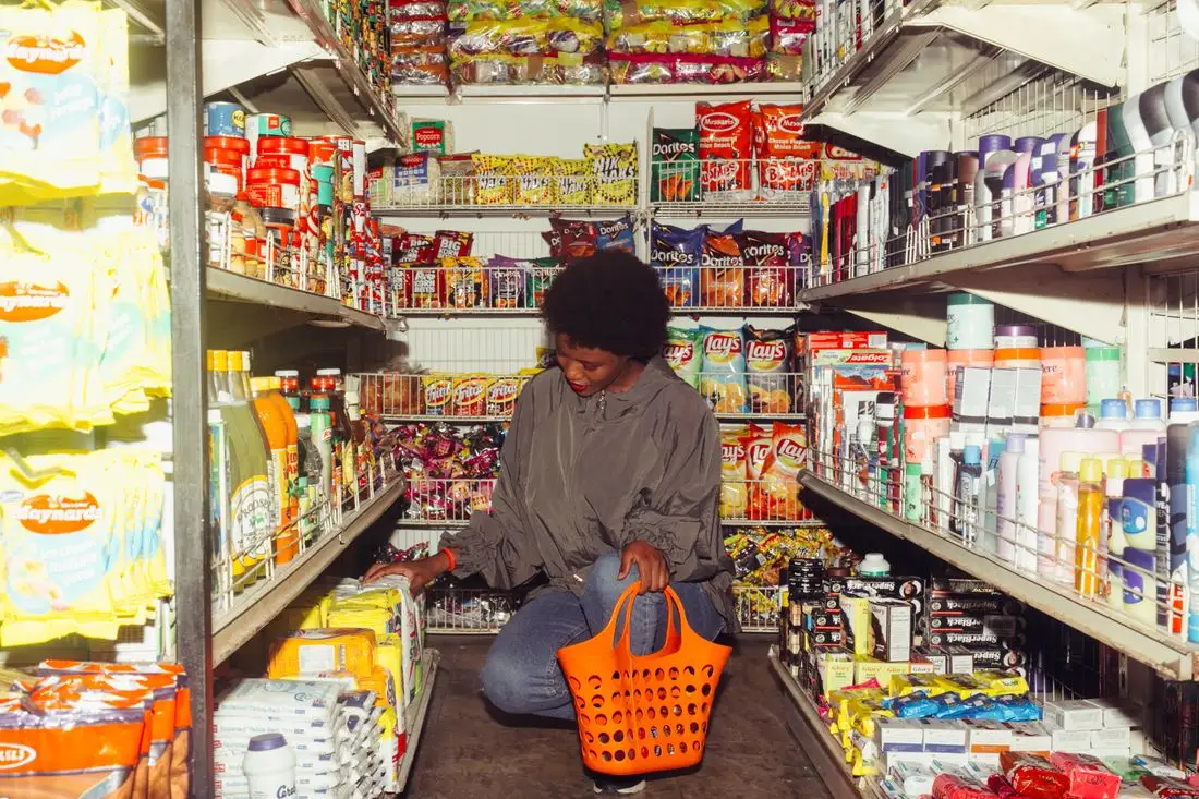 A woman crouching in a grocery aisle comparing products with an orange shopping basket beside her